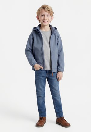 Smejoči se blond deček stoji z roko v žepu, oblečen v modro kapuco s kapuco, sivo majico, modre kavbojke in rjave škornje na belem ozadju.