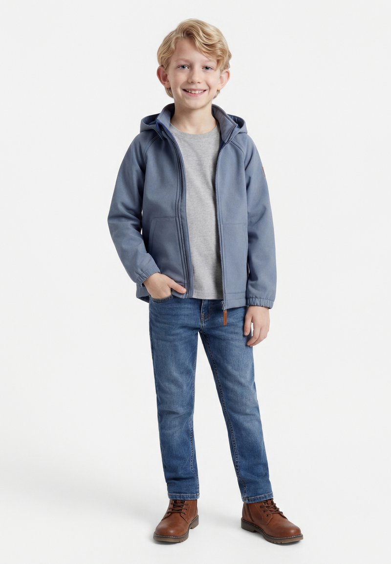 Χαμογελαστός ξανθός αγόρι που στέκεται με το χέρι στην τσέπη, φορώντας μπλε μπουφάν με κουκούλα, γκρι μπλουζάκι, μπλε τζιν και καφέ μπότες σε λευκό φόντο.