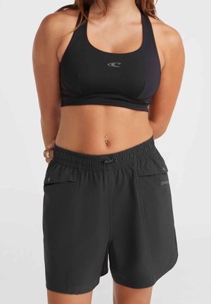 Femme portant un brassard de sport noir et un short noir ample avec une ceinture élastique, debout devant un fond clair uni.