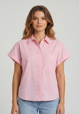 Jonge vrouw met golvend bruin haar, gekleed in een lichtroze blouse met korte mouwen en lichtblauwe spijkerbroek, staand tegen een effen achtergrond.