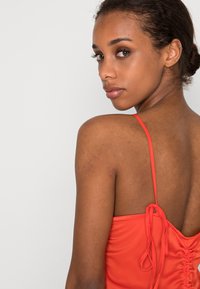 Vestido de cetim vermelho com alças finas, apresentando um detalhe atado nas costas e uma textura franzida ao longo da cintura. Acabamento liso e brilhante.