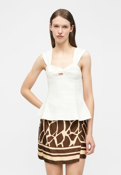 Γυναίκα με άσπρο αμάνικο τοπ με φιόγκο και καφέ φούστα με σχέδιο καμηλοπάρδαλης, στέκεται με το ένα χέρι στη μέση.