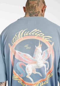 Lyseblå bomulds t-shirt med et stort trykt design af en enhjørning med vinger, omgivet af tekst og en cirkulær ramme i pastelfarver.