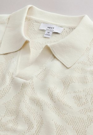 Polo en maille crème avec un motif floral texturé subtil, col plié, et une étiquette "NEXT Regular Fit" indiquant la taille M, fabriqué au Bangladesh.