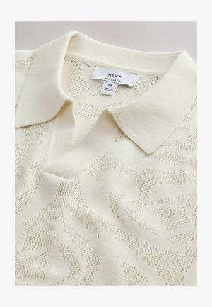 Polo in maglia crema con motivo floreale sottile e testurizzato, colletto piegato e etichetta "NEXT Regular Fit" che indica la taglia M, realizzata in Bangladesh.