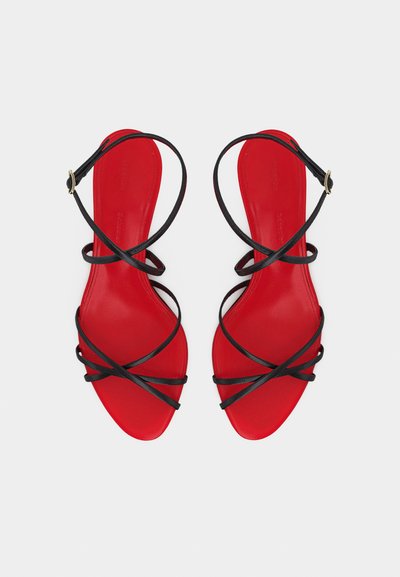 Κόκκινα φλατ σανδάλια με λεπτούς μαύρους λουριά σε σχήμα σταυρού και αγκράφες στον αστράγαλο, εμφανιζόμενα από πάνω σε ανοιχτό φόντο.