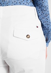 Pantaloni bianchi con tasca e bottone, dettagliati con un piccolo logo, indossati con una maglietta a maniche lunghe blu con fantasia, mostrando la vista laterale e posteriore dell'abbigliamento.