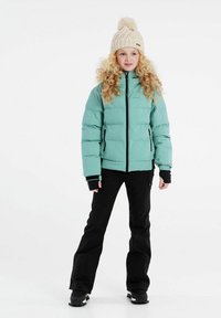Helle türkisfarbene Pufferjacke mit schwarzen Reißverschlüssen und hohem Kragen, kombiniert mit schwarzen Hosen und einer cremfarbenen Strickmütze mit einem Bommel.