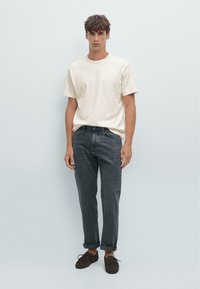 Lichtbeige T-shirt met korte mouwen gecombineerd met donkergrijze opgerolde jeans. Model draagt bruine suède veterschoenen en staat tegen een effen achtergrond.