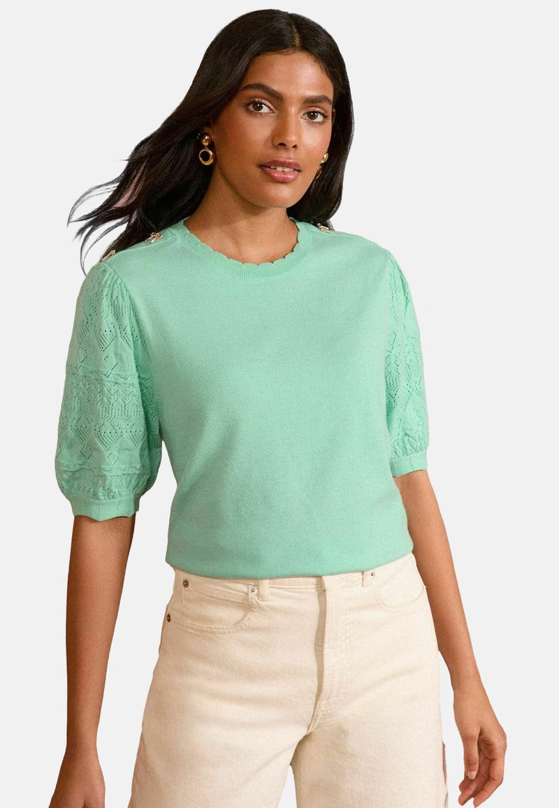 Minta zaļa īsu piedurkņu džemperis ar viļņveida dekoltē un mežģīņu raksta puff piedurknēm. Apvienots ar gaišas krāsas džinsu.