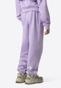 Λιλά φόρμα με ελαστική ζώνη στη μέση και στα μανσέτα, εμφανιζόμενη από την πίσω πλευρά. Διαθέτει λευκό λογότυπο στον αριστερό αστράγαλο. Φορεμένη με μπεζ αθλητικά παπούτσια.