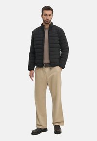 Chaqueta acolchada negra con costuras horizontales, cuello alto y cierre de cremallera. Llevada sobre un suéter beige y pantalones anchos beige.