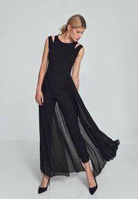 Zwarte jumpsuit met mouwloos ontwerp, featuring cut-out schouders en een vloeiende chiffon overlay. Slim fit, enkellang, aangevuld met spitse schoenen.