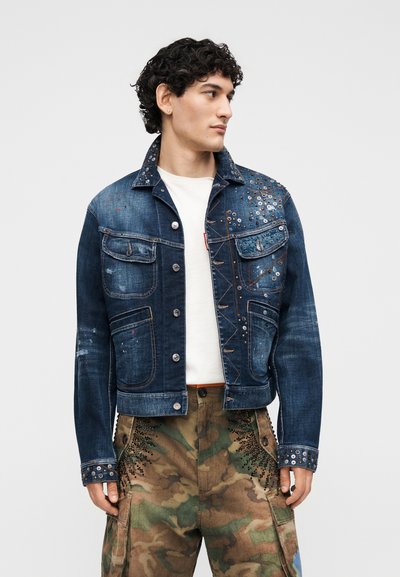 Άνδρας που φοράει σκούρο μπλε τζιν μπουφάν με τρουκς πάνω από άσπρο πουκάμισο και παντελόνι καμουφλάζ cargo με μαύρες διακοσμητικές χάντρες, κοιτάει στο πλάι.