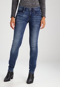 Jeans oscuros de corte slim, hechos de mezclilla. Con bolsillos frontales y cierre de botón. Combinados con botas negras de tacón.