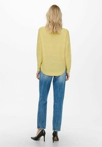 Gelb gestreifter Pullover mit abgerundetem Saum, kombiniert mit hellblauen, ausgewaschenen Jeans und schwarzen High-Heels. Von hinten betrachtet.
