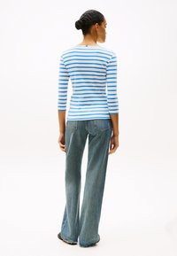 Μπλε και λευκή ριγωτή μακρυμάνικη μπλούζα που φαίνεται από πίσω, συνδυασμένη με ανοιχτόχρωμα μπλε καμπάνα τζιν και μαύρα παπούτσια, σε ουδέτερο φόντο.