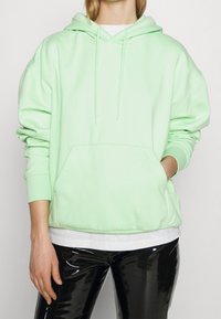 Sudadera con capucha verde menta con un bolsillo frontal y cordones, combinada con leggings negros brillantes. Tejido suave, corte relajado.