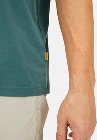 Teal bomulds T-shirt med løs pasform, der har et lille gult logo tag på venstre side. Korte ærmer og en rund halsudskæring.