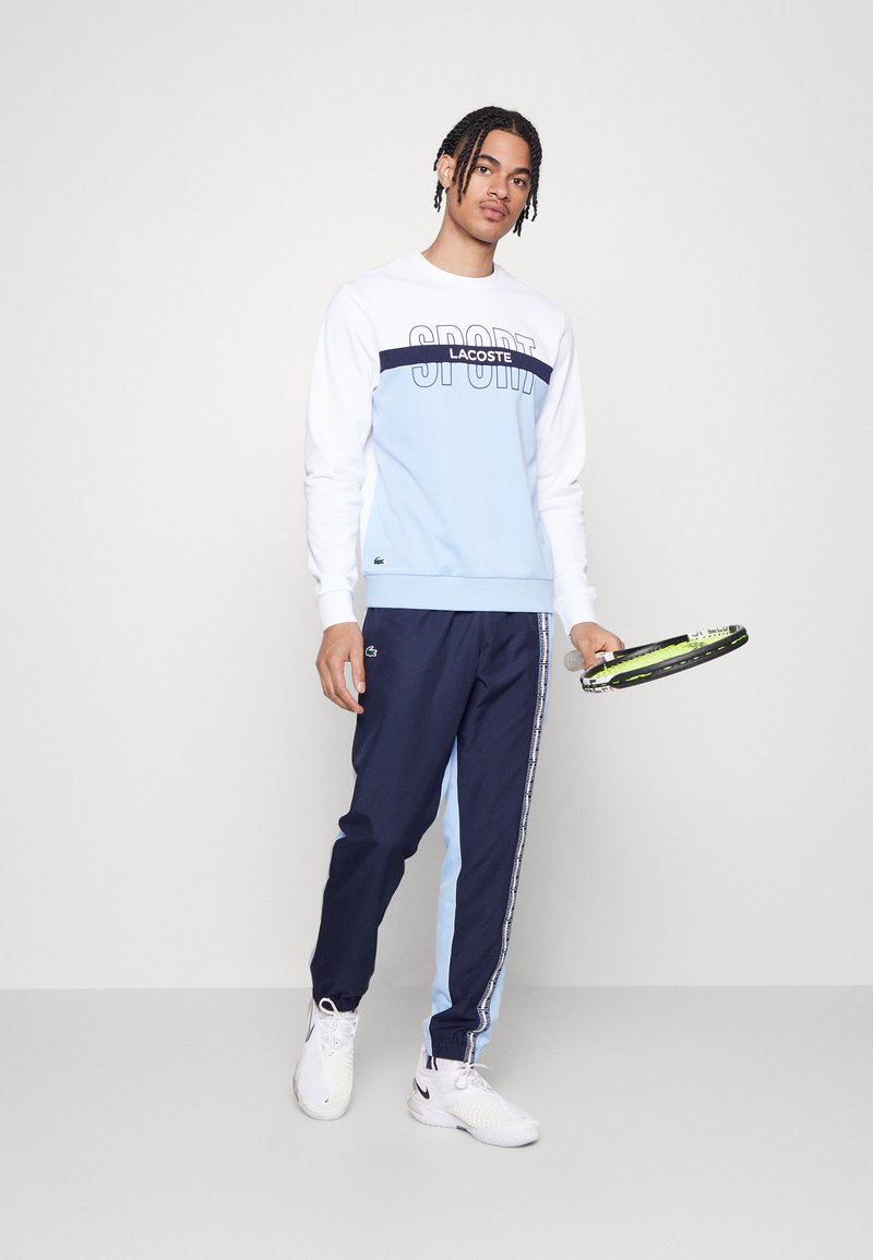 Sudadera deportiva en blanco y azul claro con una franja negra, con el logo de "LACOSTE"; combinada con pantalones de chándal navy con paneles laterales en azul claro.