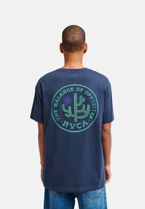 Uomo con capelli corti che indossa una t-shirt blu navy con un cactus e la scritta "The Balance of Opposites RVCA" sulla schiena, abbinata a jeans blu.