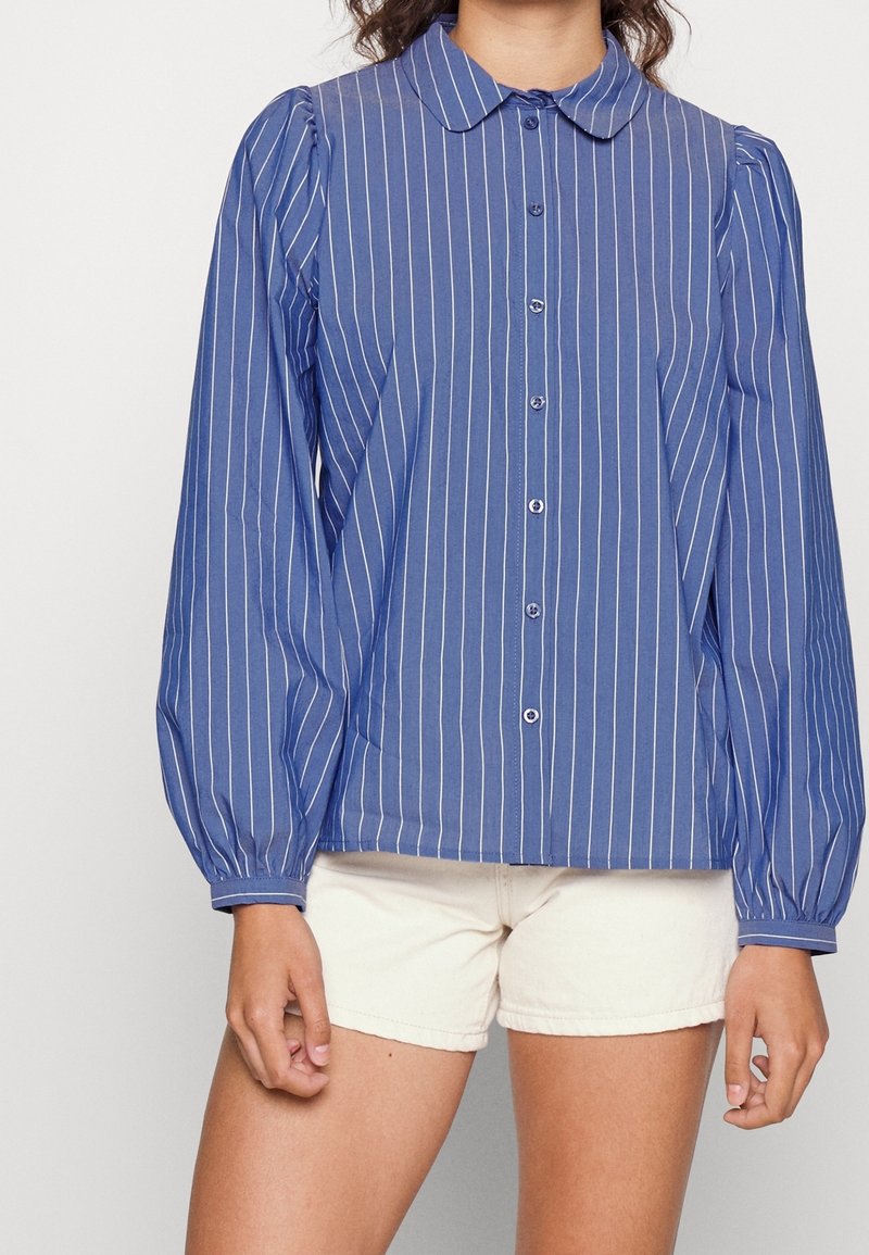 Femme portant une chemise bleue à manches longues avec des rayures blanches verticales et un short blanc, debout devant un fond uni.