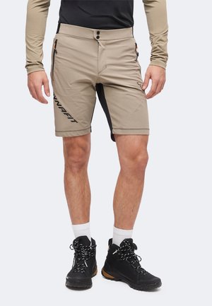 Mann trägt beige Outdoor-Shorts mit seitlichen Reißverschlusstaschen, schwarze Wanderschuhe, weiße Socken und ein langärmeliges Hemd mit schwarzen und beigen Ärmeln.