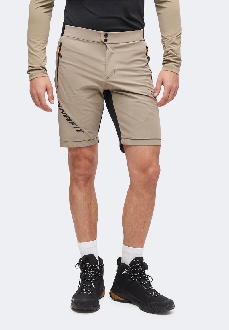 Homme portant un short beige de plein air avec poches latérales zippées, des chaussures de randonnée noires, des chaussettes blanches et une chemise à manches longues avec des manches noires et beiges.