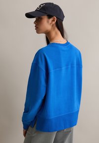 Niebieska, oversize'owa bluza z prążkowanymi mankietami i dołem. Dobraną do niej czarna czapka z białym logo oraz srebrny łańcuszek na szyi.