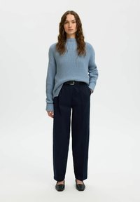 Jasnoniebieski sweter z dzianiny o prążkowanej fakturze, zestawiony z granatowymi spodniami o szerokich nogawkach i czarnymi płaskimi butami. Modelka stoi na tle jednolitego tła.