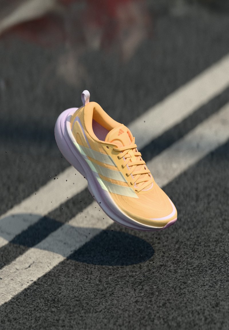 Scarpa da corsa arancione chiaro con strisce bianche sospesa in aria sopra una strada asfaltata segnata da linee bianche, con piccole particelle nere che si disperdono.