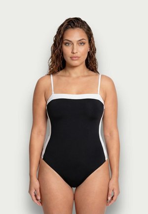 Donna con capelli mossi che indossa un costume intero nero con rifinitura bianca e sottili spalline, in piedi davanti a uno sfondo chiaro uniforme.