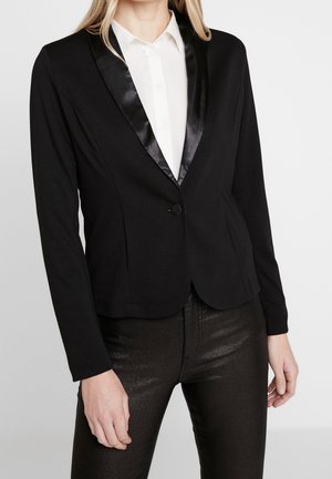 Kobieta ubrana w dopasowaną czarną marynarkę z satynowymi klapami, białą koszulę zapinaną na guziki i ciemne teksturowane spodnie, stojąca na tle jednolitego tła.