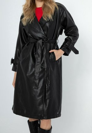 Mujer con un abrigo trench negro de cuero con cinturón sobre una camiseta roja, con tiras con hebillas en las mangas y botas negras acolchadas visibles debajo del abrigo.