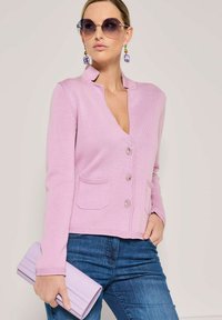 Frau in einem lavendelblauen, geknöpften Cardigan und blauen Jeans, die eine passende lavendelblaue Clutch hält, accessoriert mit großen lila Ohrringen und Sonnenbrille.