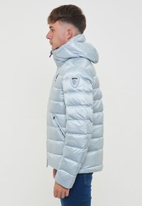 Giacca a vento azzurro chiaro con cappuccio, caratterizzata da un design trapuntato, tasche con zip e una toppa con logo sulla manica sinistra.