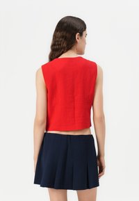 Top corto sin mangas de color rojo en tejido texturizado con dobladillo recto. Combinado con una falda azul marino con pliegues, que presenta un diseño minimalista.