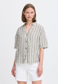 Camicia a maniche corte con bottoni di colore beige e strisce verticali nere, con colletto e maniche arrotolate. Indossata con pantaloni corti bianchi.