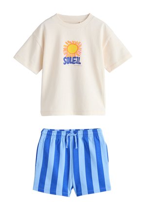 Cremefarvet kortærmet skjorte med solmotiv og tekst "SOLEIL" stylet sammen med blå elastiske shorts med lodrette striber og snøre i taljen.