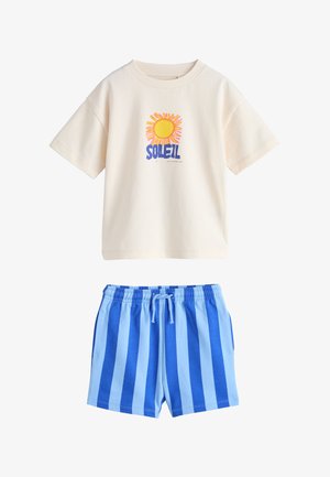 Cremefarvet kortærmet skjorte med solmotiv og tekst "SOLEIL" stylet sammen med blå elastiske shorts med lodrette striber og snøre i taljen.