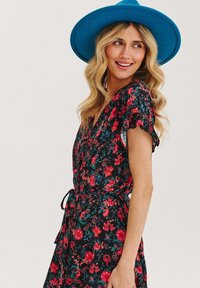 Květinové zavinovací šaty z tmavé látky s červenými a modrými květinami. Krátké volánkové rukávy, zavazování v pase a výstřih do V. Doplněno modrým kloboukem s širokým krempěm.