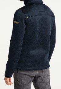 Granatowa polarowa kurtka z wysokim kołnierzem, o teksturowanym wykończeniu, subtelnym wzorze i żółtymi akcentami na ramionach. Noszona z szarymi dżinsami.