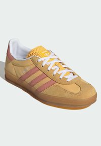 Sneaker Adidas Gazelle in suede giallo, con strisce rosa, lacci bianchi e una linguetta gialla texturizzata con logo. Suola in gomma visibile sotto.
