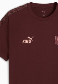 Camiseta color marrón burdeos con cuello redondo, mangas cortas, franjas verticales en los hombros y logotipos impresos de "KING" y Puma en el pecho.
