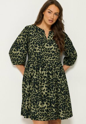 Kvinde med langt brunt hår iført en grøn og sort leopardprint kjole med trekvartlange pufærmer mod en ensfarvet lys baggrund.