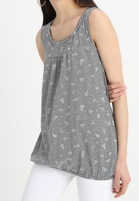 Kvinna i ärmlös svart-vit randig blus med små ankarmönster och vita byxor, håller armen över midjan.