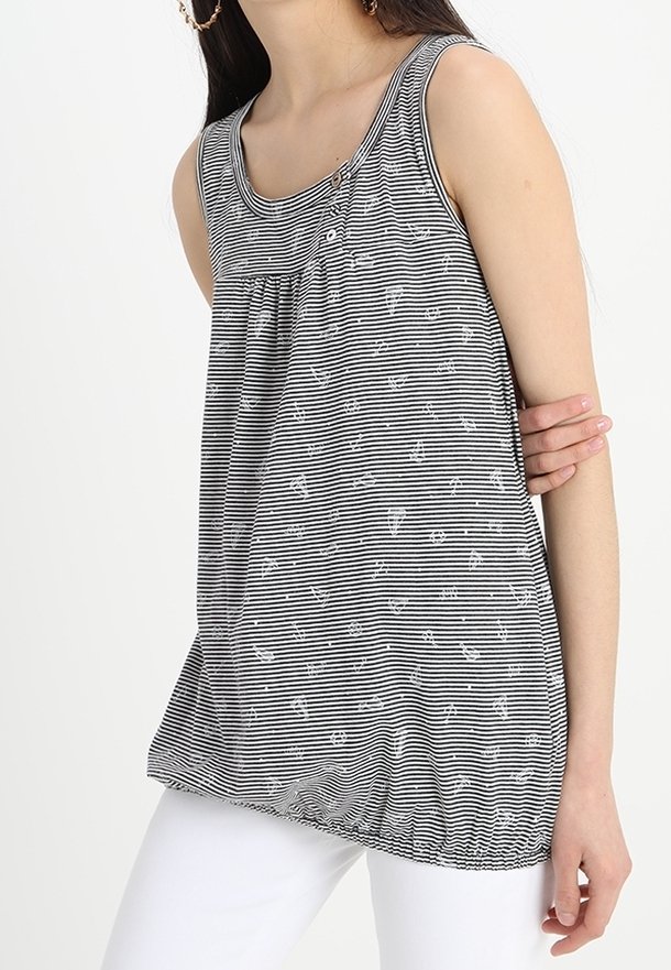 Kvinna i ärmlös svart-vit randig blus med små ankarmönster och vita byxor, håller armen över midjan.