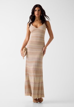 Frau trägt ein ärmelloses, gestricktes Maxikleid in Beige und Pastellstreifen, hält eine mit Fransen verzierte Clutch und trägt goldene Creolen sowie Sandalen.