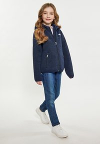 Jūros karininkų zip-up striukė, pagaminta iš struktūrinio audinio, turi gobtuvą ir dvi užtrauktuko kišenes. Dėvima su mėlynais džinsais ir baltais sportbačiais.