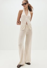 Beige stickad tvådelad set som har en halterneck-top med knappar framtill och lösa, utsvängda byxor. Strukturerad textil och snörningsdetalj.